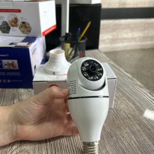 Mano sosteniendo una cámara de seguridad inteligente con diseño de foco blanco sobre un escritorio.