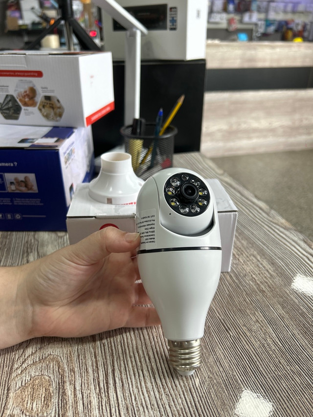 Mano sosteniendo una cámara de seguridad inteligente con diseño de foco blanco sobre un escritorio.
