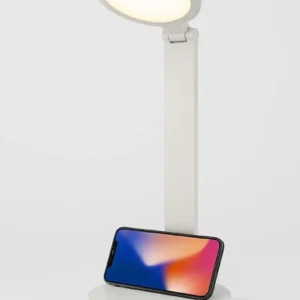 Lámpara de escritorio blanca con luz encendida y un smartphone posicionado horizontalmente en su base circular.